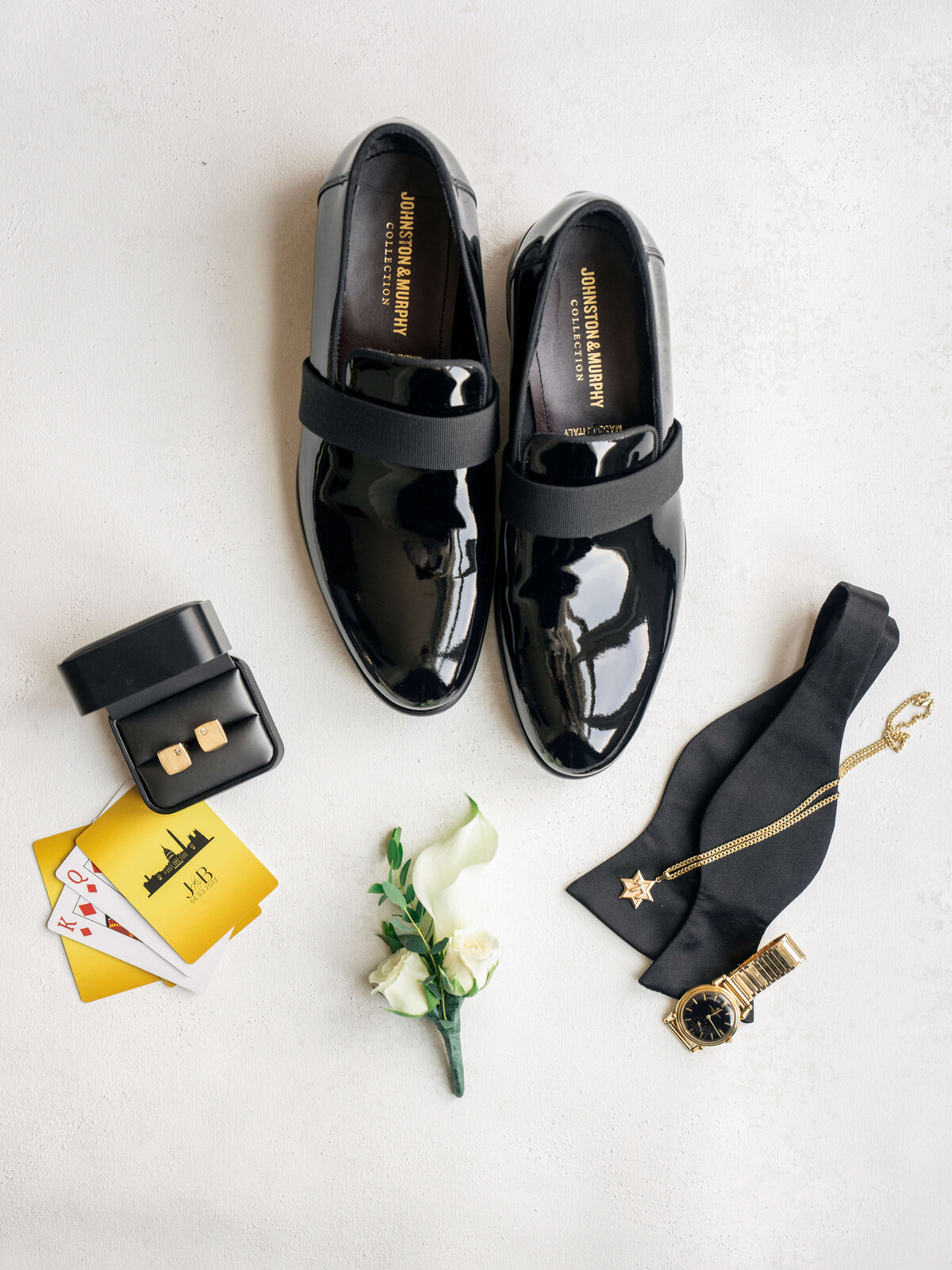 A pair of glossy black shoes, a black bow tie, gold watch, star necklace, cufflinks in a box, a white flower, and playing cards with a yellow card on a light surface.