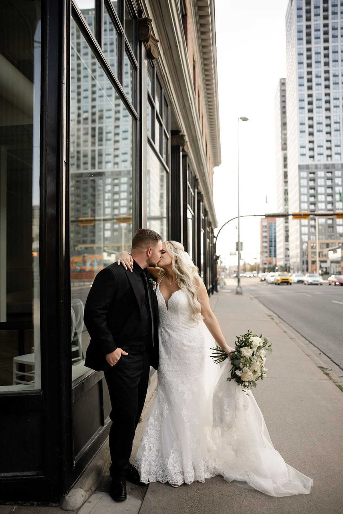 A newlywed couple shares a romantic kiss outside Studios at Vic Park in downtown Calgary. The bride, in a lace wedding gown, holds a bouquet of white flowers, while the groom, dressed in a black suit, leans against the building. The urban backdrop features modern high-rises and city streets, blending elegance with a chic downtown vibe.