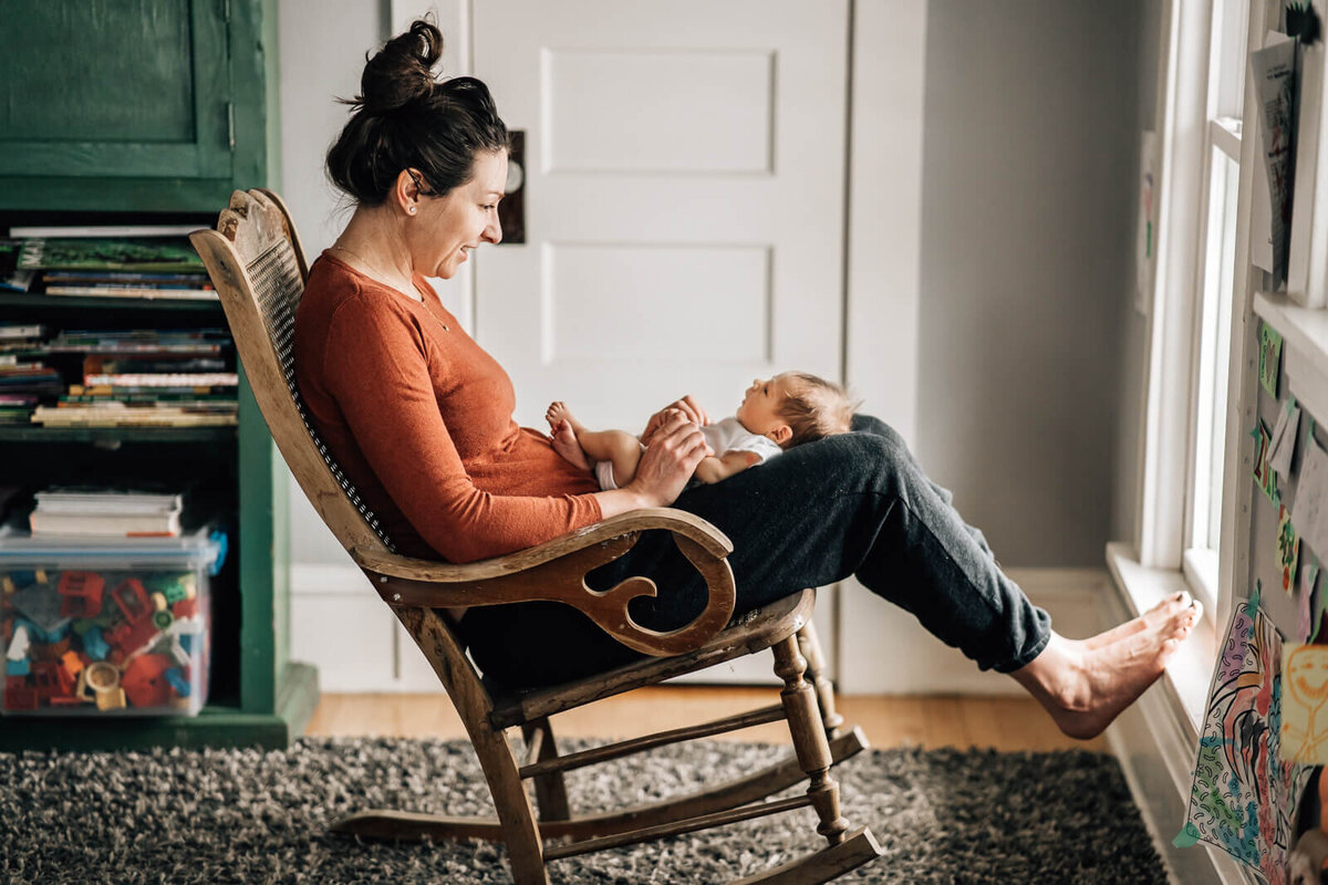 A mother sits in a rocking chair, looking down at her newborn baby on her lap during an in home newborn session with Kate Simpson.