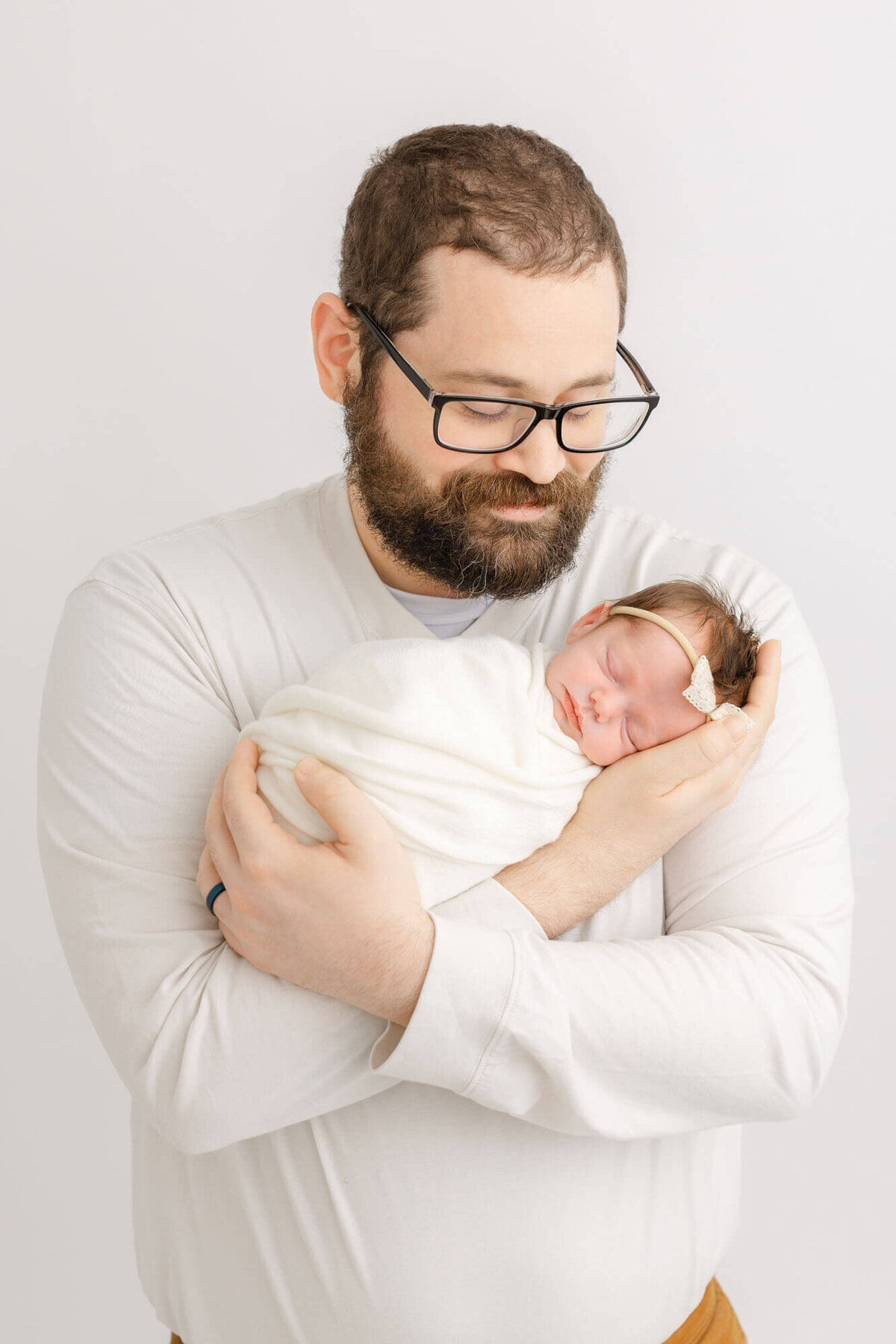 New Dad holding baby in his arms and smiling down at her. He is dressed in white and baby is swaddled in white and wearing a minimalist white headband.
