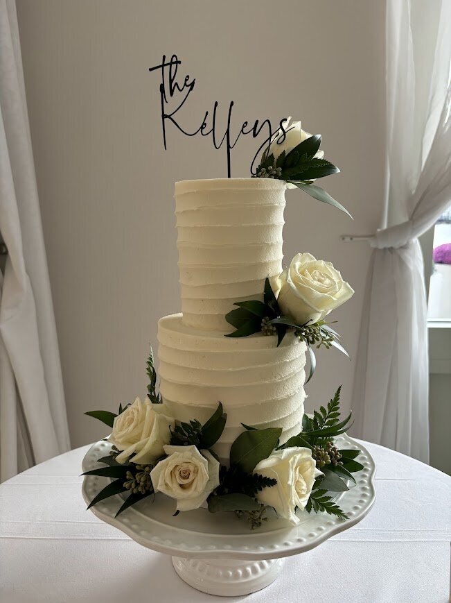 A small two tier cake is pictured on a white cake stand. The cake has line textured white buttercream and is decorated with fresh greenery and white flowers.