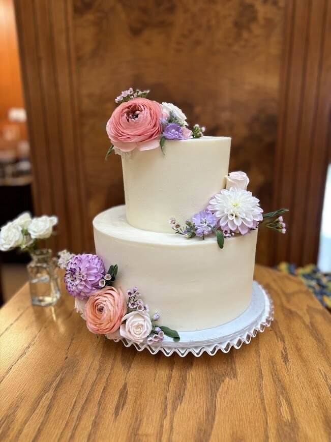 A two tier cutting cake sits on a wood table. The frosting is smooth white, and there are clusters of spring flowers in pastel pinks and purples.