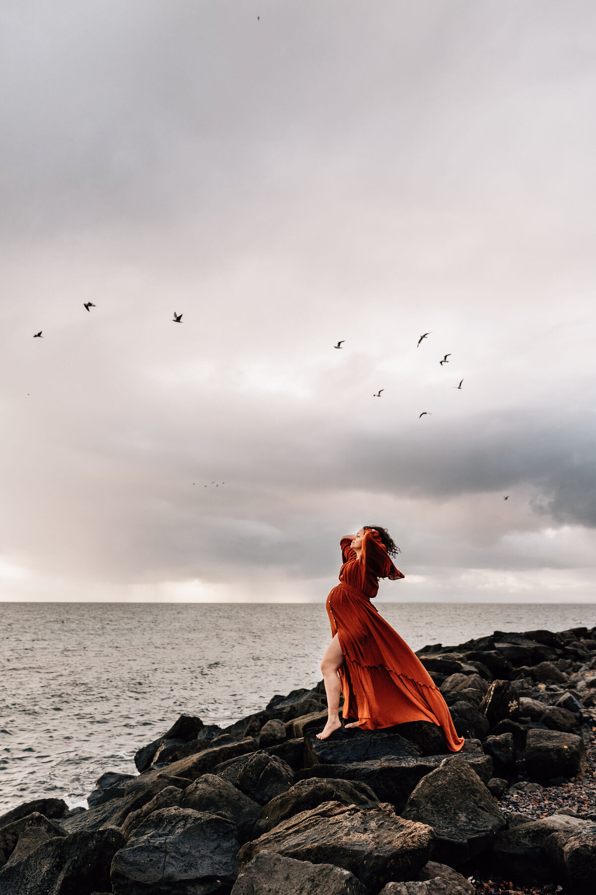 Pregnant woman stands on beach rocks with flock of birds flying overhead
