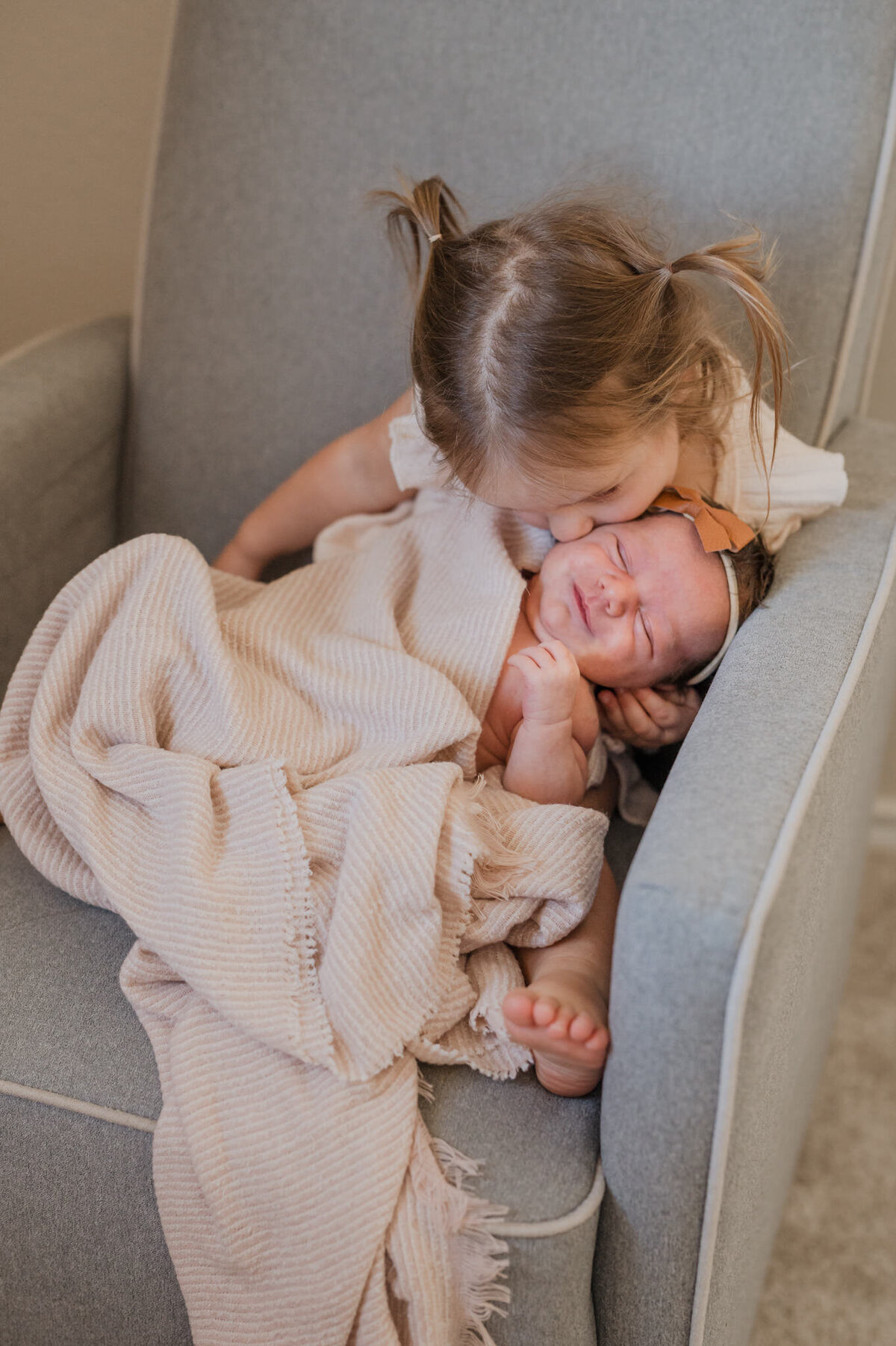 Toddler kisses her baby sister on the cheek while they sit together in the nursery rocker, taken by San Antonio newborn photographer Cassey Golden.