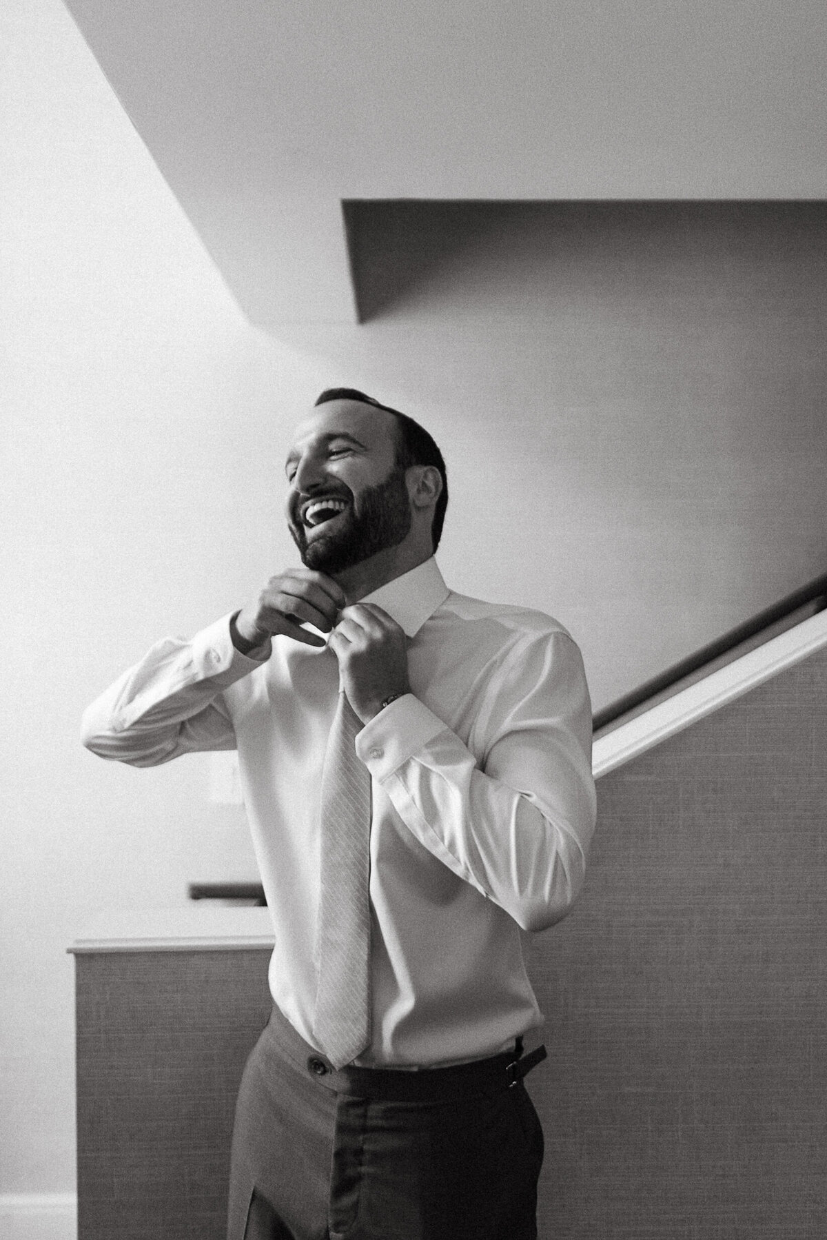 Black-and-white portrait of a groom tying his own tie.