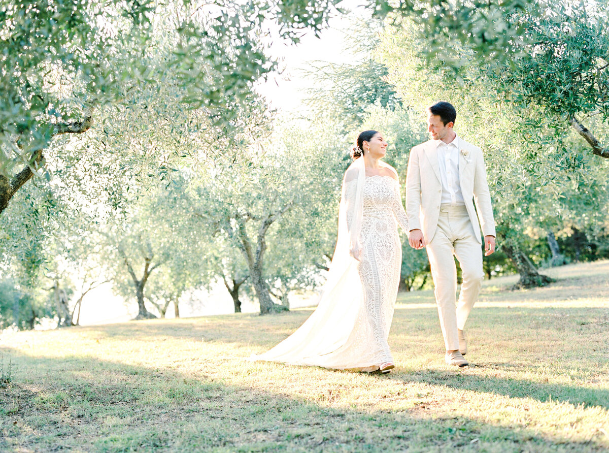 Film photograph of bride in all lace wedding dress with long veil walking with groom through olive trees photographed by Italy wedding photographer at Villa Montanare Tuscany wedding