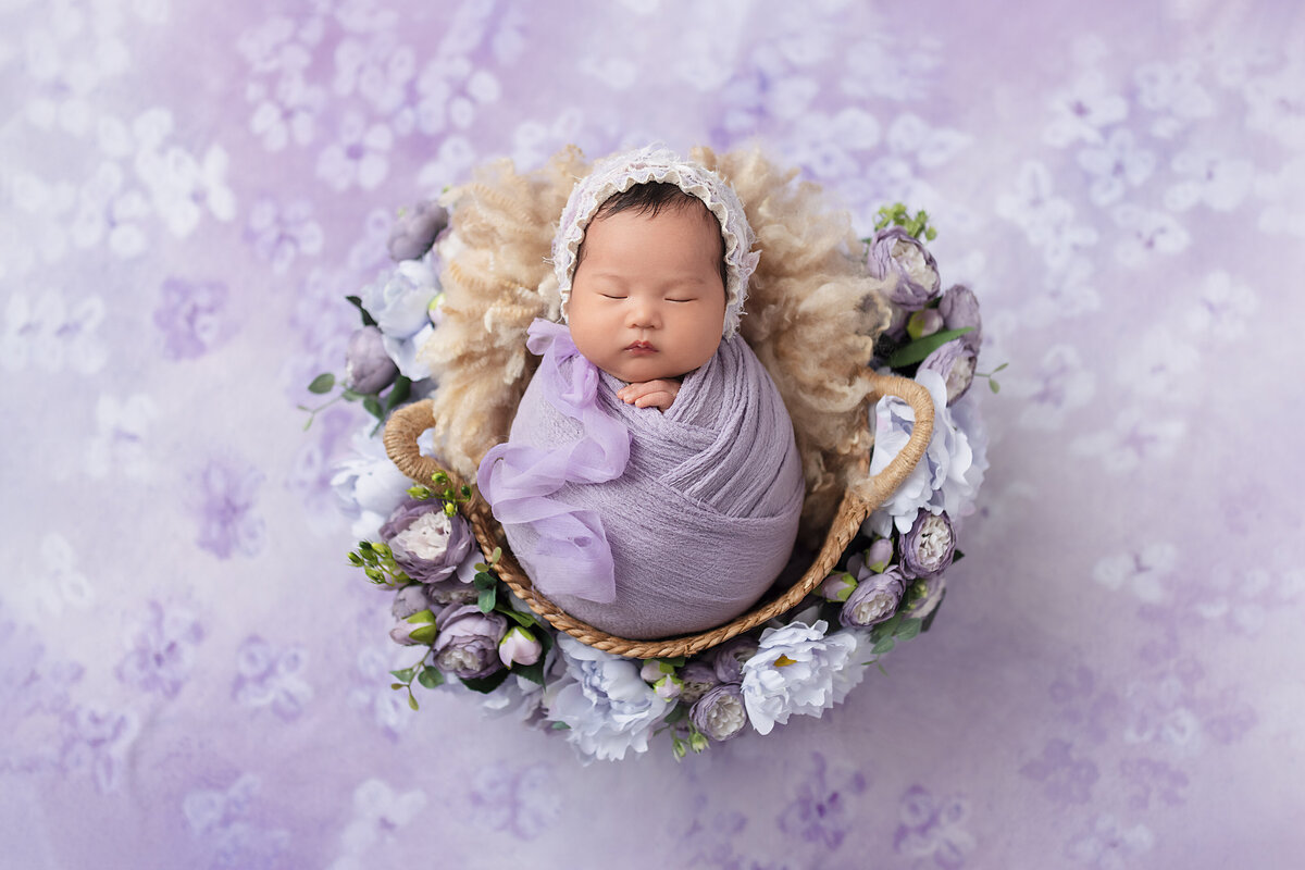 Newborn baby girl wrapped in lavender posed in a floral basket with purple blooms.