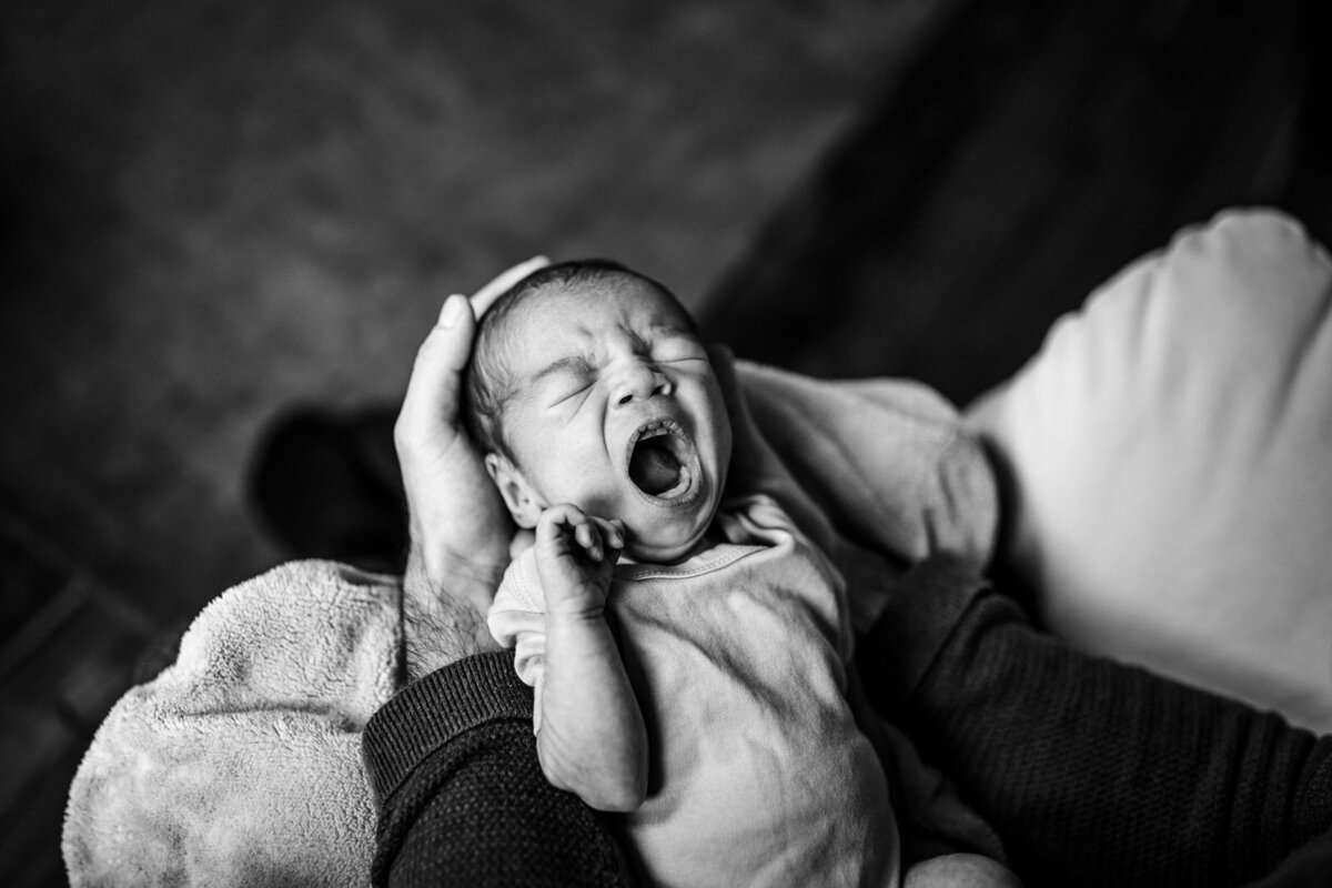 yawning newborn laying in dad's arms with his hands around baby's head