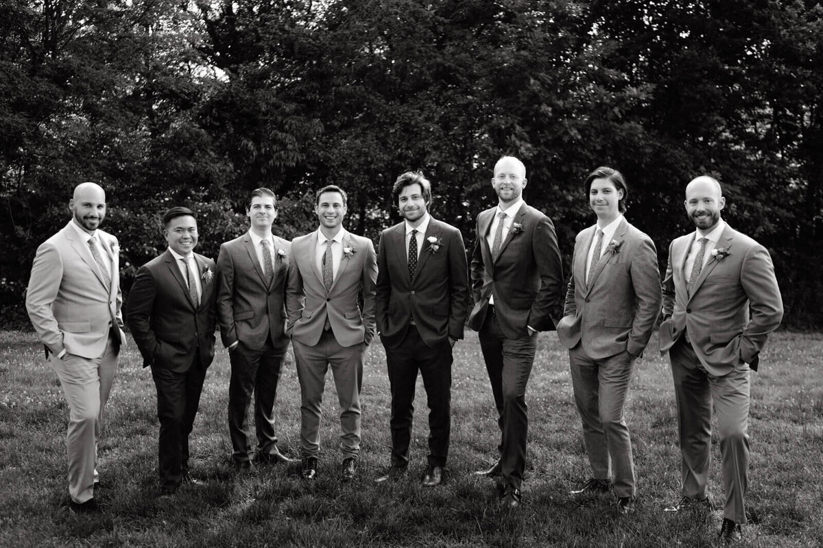 Eight men, standing, wearing suits and ties, with trees in the background