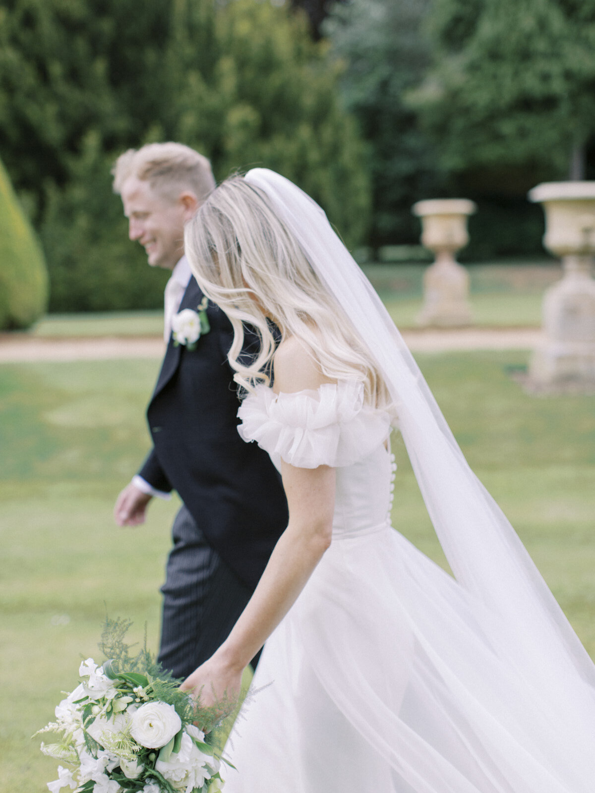 bride and groom relaxed moment walking in the gardens, bride holding white flower bouquet and groom smiling