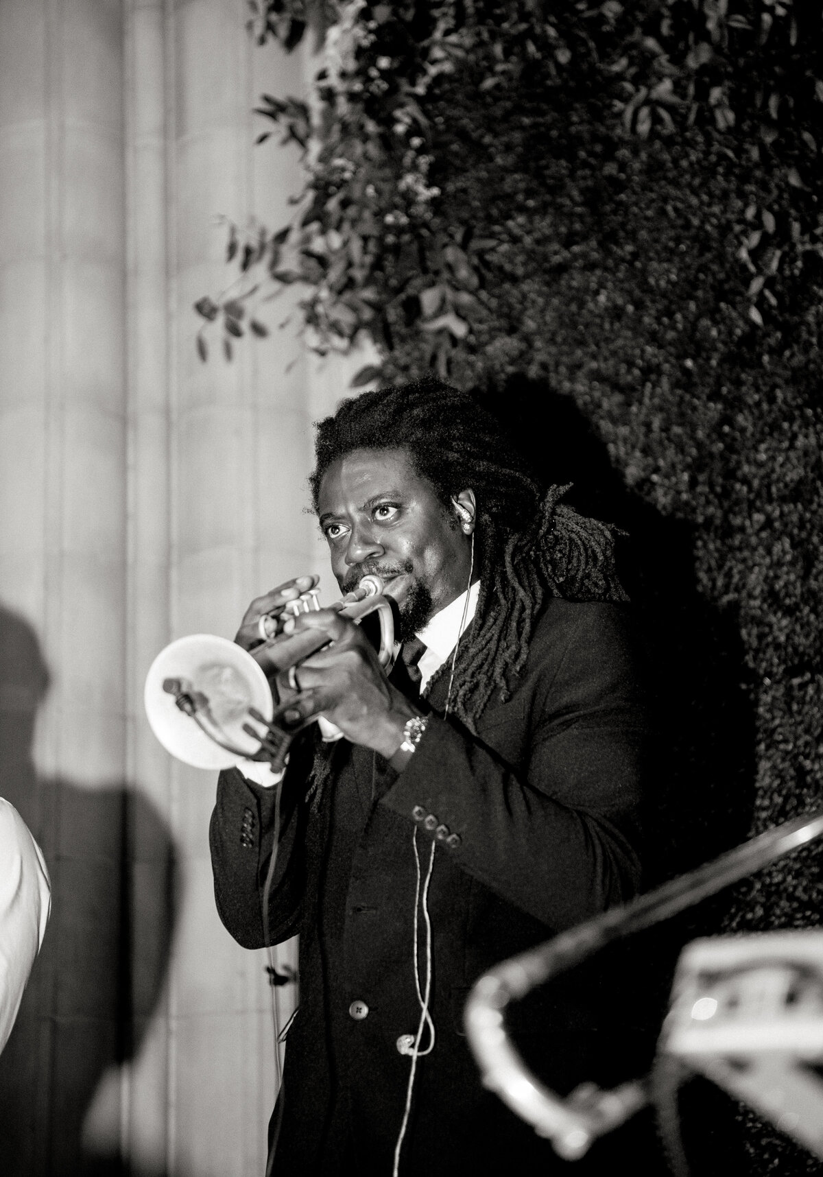 The photo was captured at Washington D.C., particularly at the Washington National Cathedral, by photographer Twah Dougherty. A musician with dreadlocks, attired in a suit, is depicted playing the trumpet with remarkable concentration and intensity. The background showcases foliage and a column, suggesting an outdoor or garden setting. This striking image, presented in black and white, amplifies the focal point on the musician, infusing a timeless quality into the scene. Additionally, Current Films is recognized as the videographers for this project.
