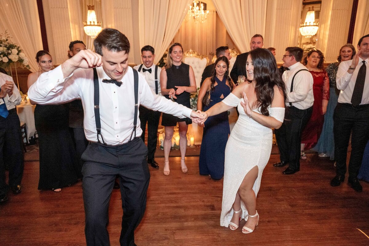 A couple, dressed formally, dances joyfully at a wedding reception. The man wears a white shirt, black pants, and suspenders, while the woman is in a white dress. Guests in evening attire surround them, smiling and clapping in celebration.