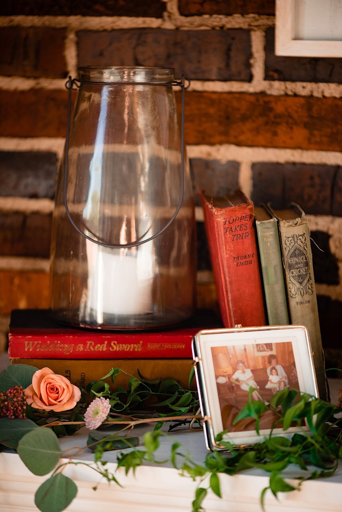 Sitting in front of an exposed brick wall is a shelf with antique books and a large clear glass hurricane lamp with a white pillar candle. In front of the lantern is a n old photograph with greenery and a peach tea rose.