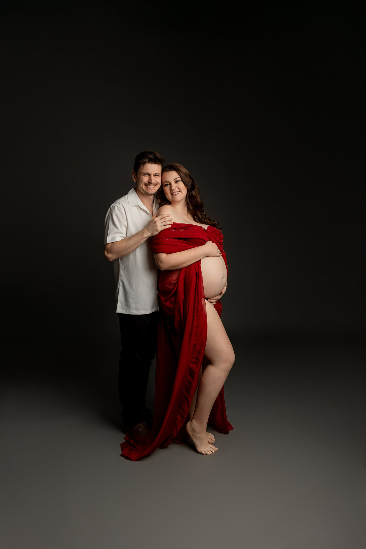 The expectant mother, wearing a red dress, stands with her husband in a side pose against a dark backdrop. Captured by Inland Empire maternity photographer Kaitlyn Dawn Photography.