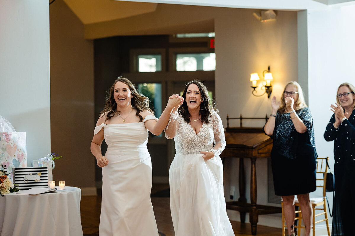 The brides entering the reception hall, holding hands and laughing joyfully, with guests clapping in the background.