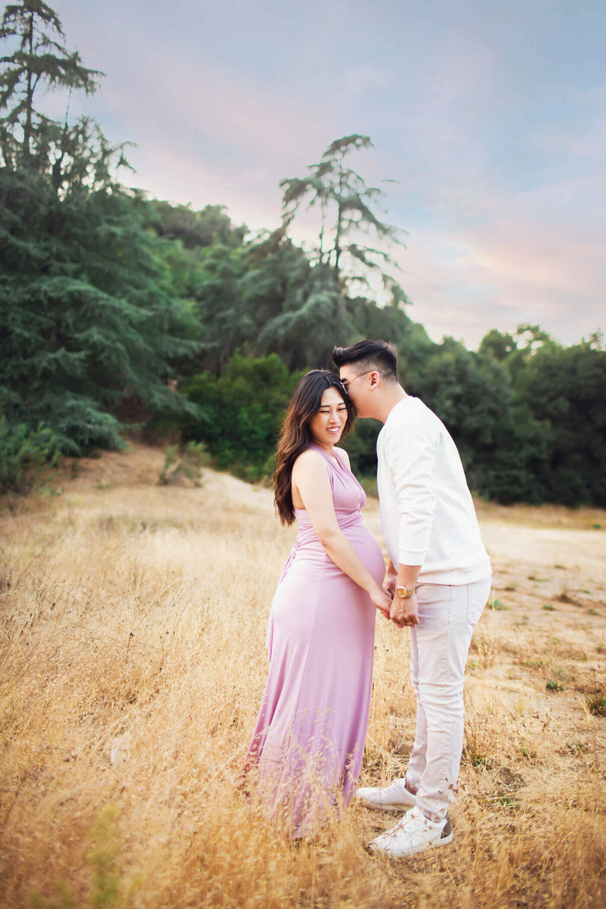 A pregnant woman stands in a golden field, holding hands with her partner. She is wearing a form-fitting, sleeveless lavender dress that accentuates her baby bump. Her partner, dressed in a white long-sleeve shirt and light pants, leans in to kiss her forehead. The couple shares a tender moment, with greenery and a colorful sky in the background, creating a serene and loving atmosphere.