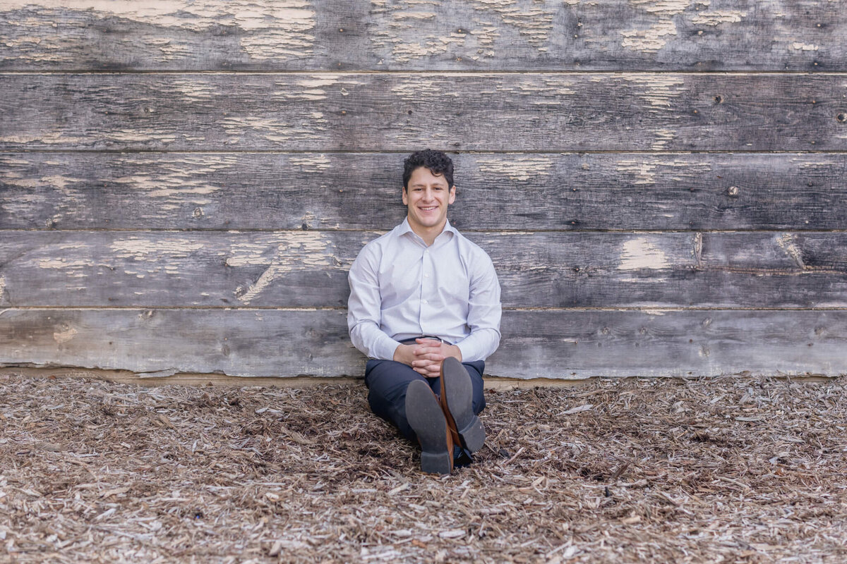 A young man with dark hair sitting in wood chips with his feet crossed at the ankles, leaning against a grey and beige wooden wall, looking at the camera from afar. Captured in Irvine.