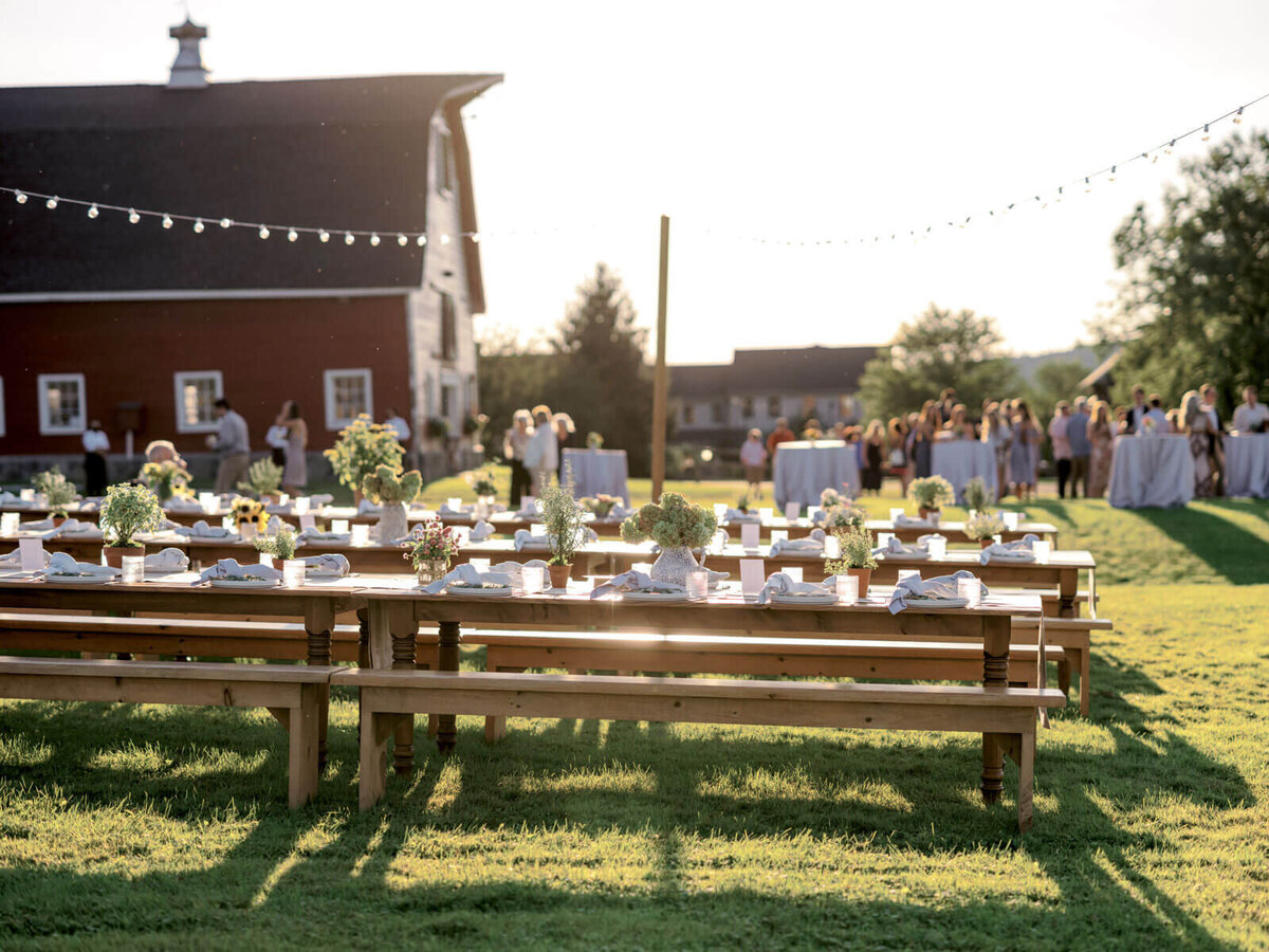 Rustic dining tables are set up outdoors, with the guests having cocktails and a big barn in the background at Lion Rock Farms, CT.