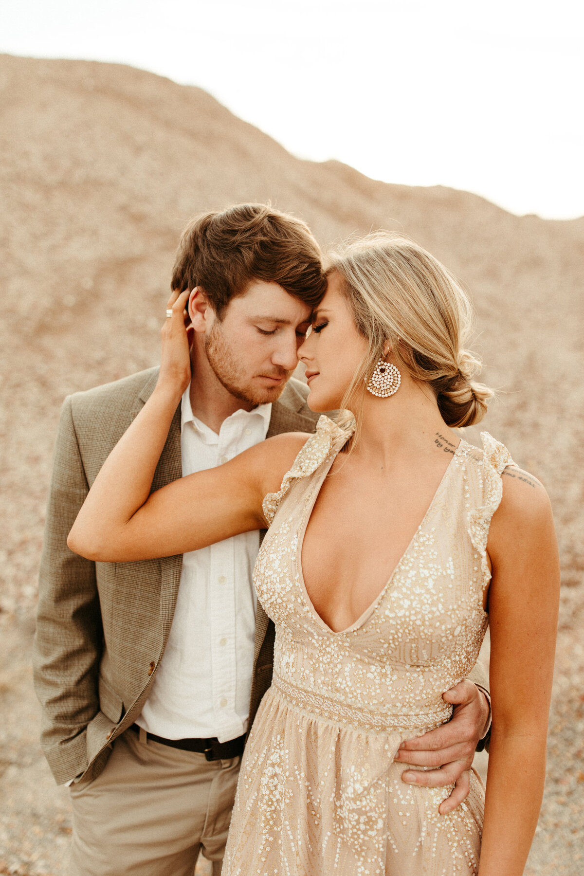 A girl in a sparkly champagne colored dress and a messy updo is standing in front of her fiancé while she pulls his face in close to hers.