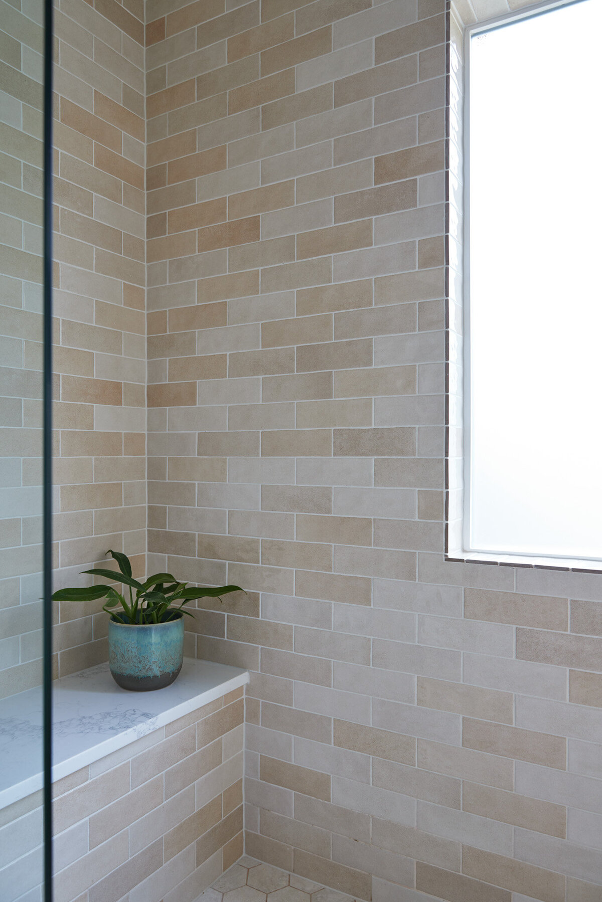 A shower with brick set subway tile that’s beige and salmon in tone. A window in the right hand corner of the shower. A shower bench with a white marble top and a plant sitting on it in a blue vase.