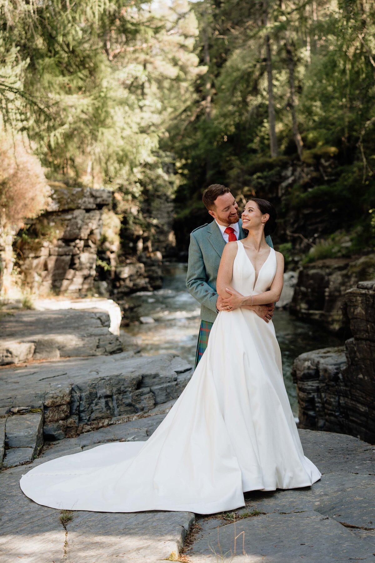 A couple are photographed with their arms wrapped around each other. They stand on rocks in a wide river setting.