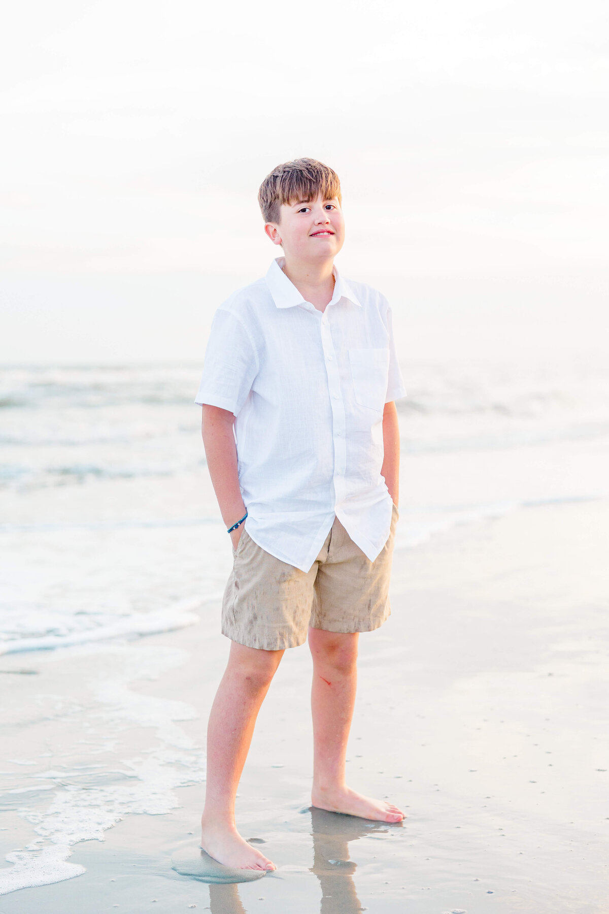 A boy stands on a sandy beach wearing a white shirt and khaki shorts. He has his hands in his pockets and looks ahead. The ocean waves are visible in the background under a light, cloudy sky.