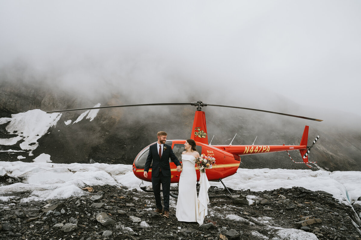 couple in wedding tire holding hands smiling at each other in front of a red helicopter and foggy mountains on their elopement day