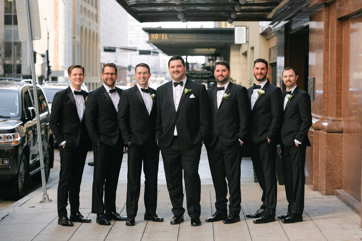 portrait of the groom and groomsmen standing outside the entrance of The Adolphus in Dallas, captured in a classic and elegant wedding group photo.