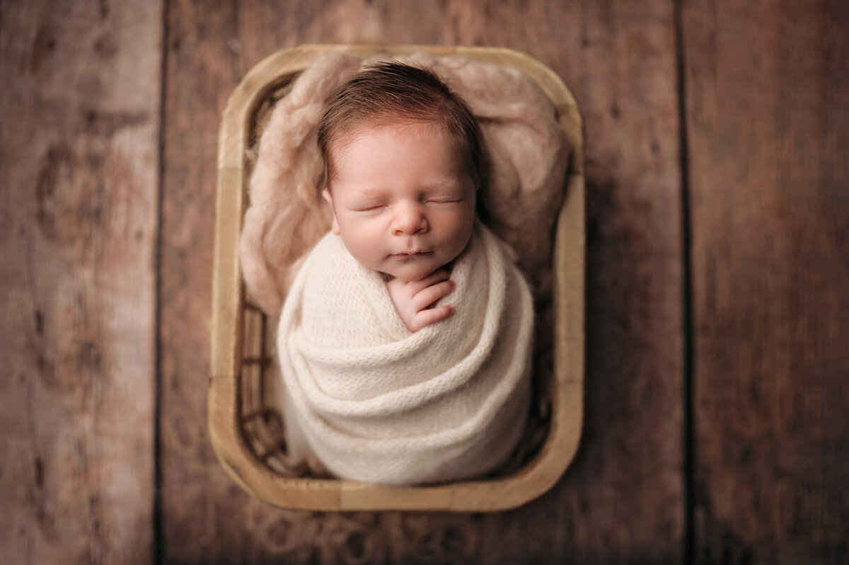 red haired newborn baby boy swaddled in cream wrap and posed inside of rectangular wooden basket on wooden floor