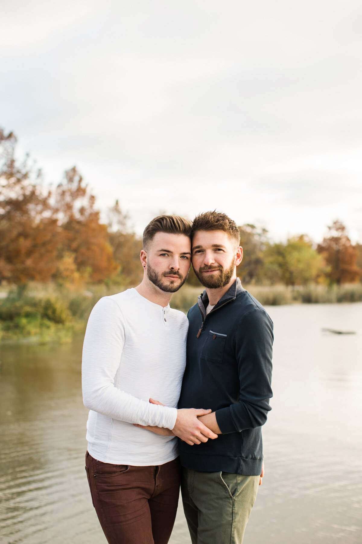 A gay couple holding each other close in front of a pond and a wooded background during their outdoor engagement session in Dallas, Texas. The man on the right is wearing a navy jacket with green pants while the man on the left is wearing a long-sleeve white shirt with brown pants.