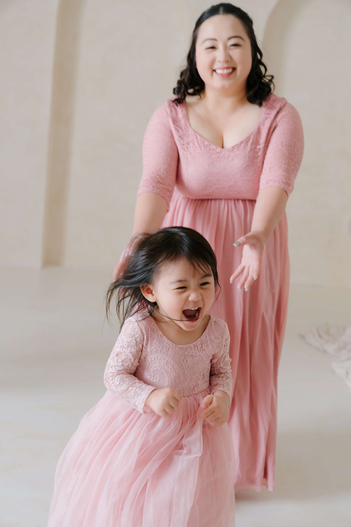 A mother and her little girl, both in elegant pink dresses, delight in a playful chase.