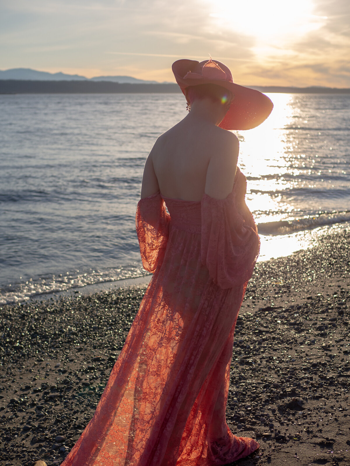 A pregnant person weaering a pink, lacy dress walks along the Sound at sunset in Edmonds, WA.