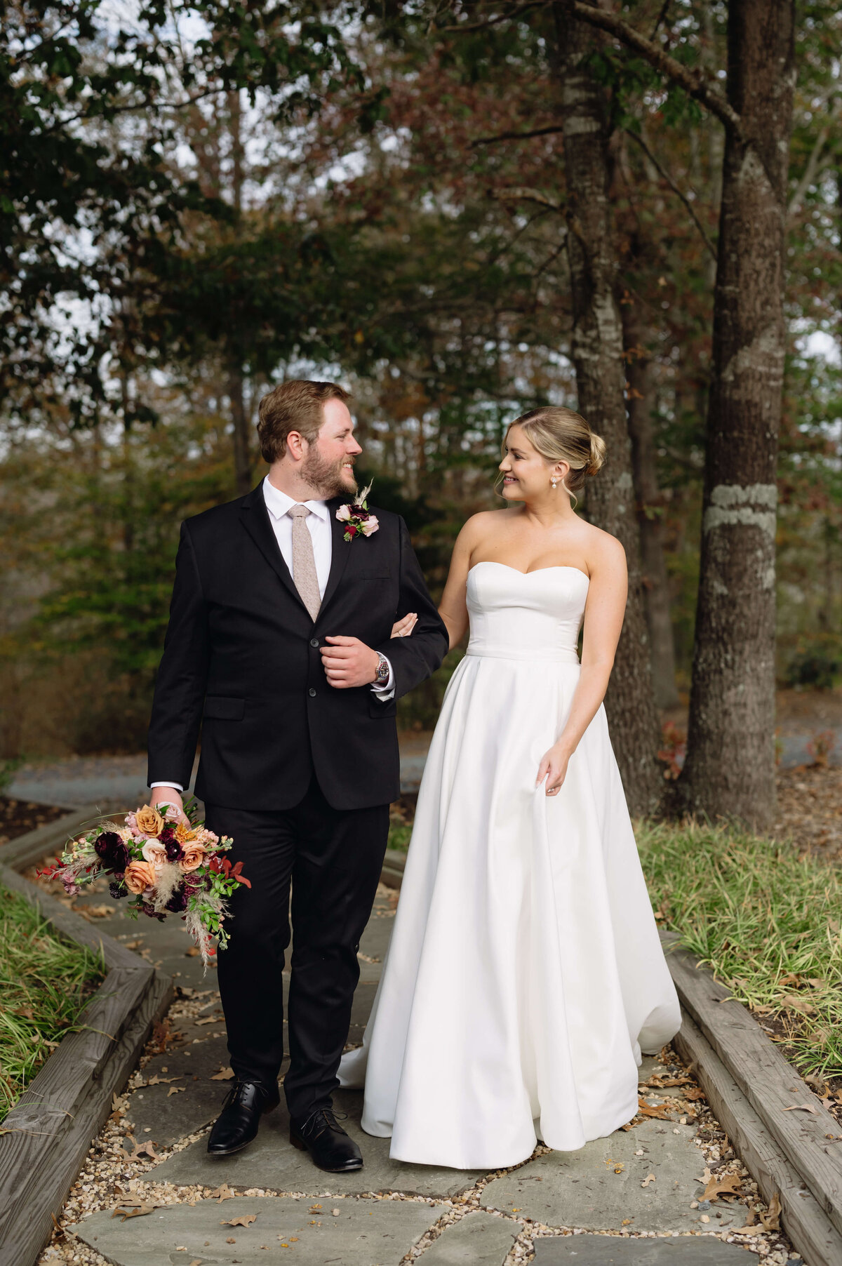 outdoor wedding picture with bride holding her grooms arm while they look at each other and smile while on a stone trail in the woods near Charlottesville wedding venues lawn