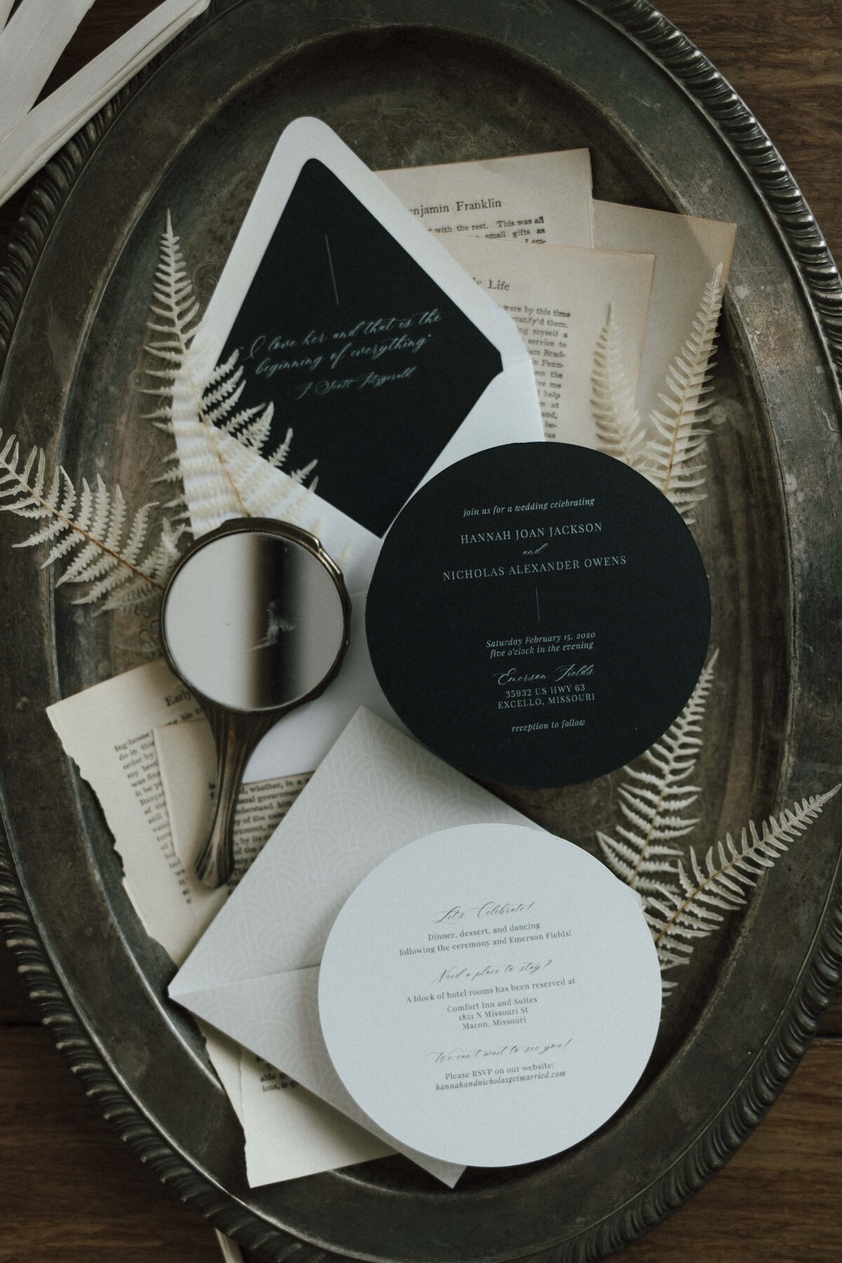 Rounded white and dark green wedding invitations with white cursive font atop a weathered silver tray with loose book pages.