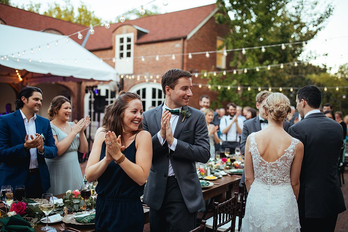 guests cheering for the bride in a lace dress and groom in a navy suit as they enter the wedding reception at cheekwood botanical gardens with string lighting and exposed brick at the Frist Learning Center.