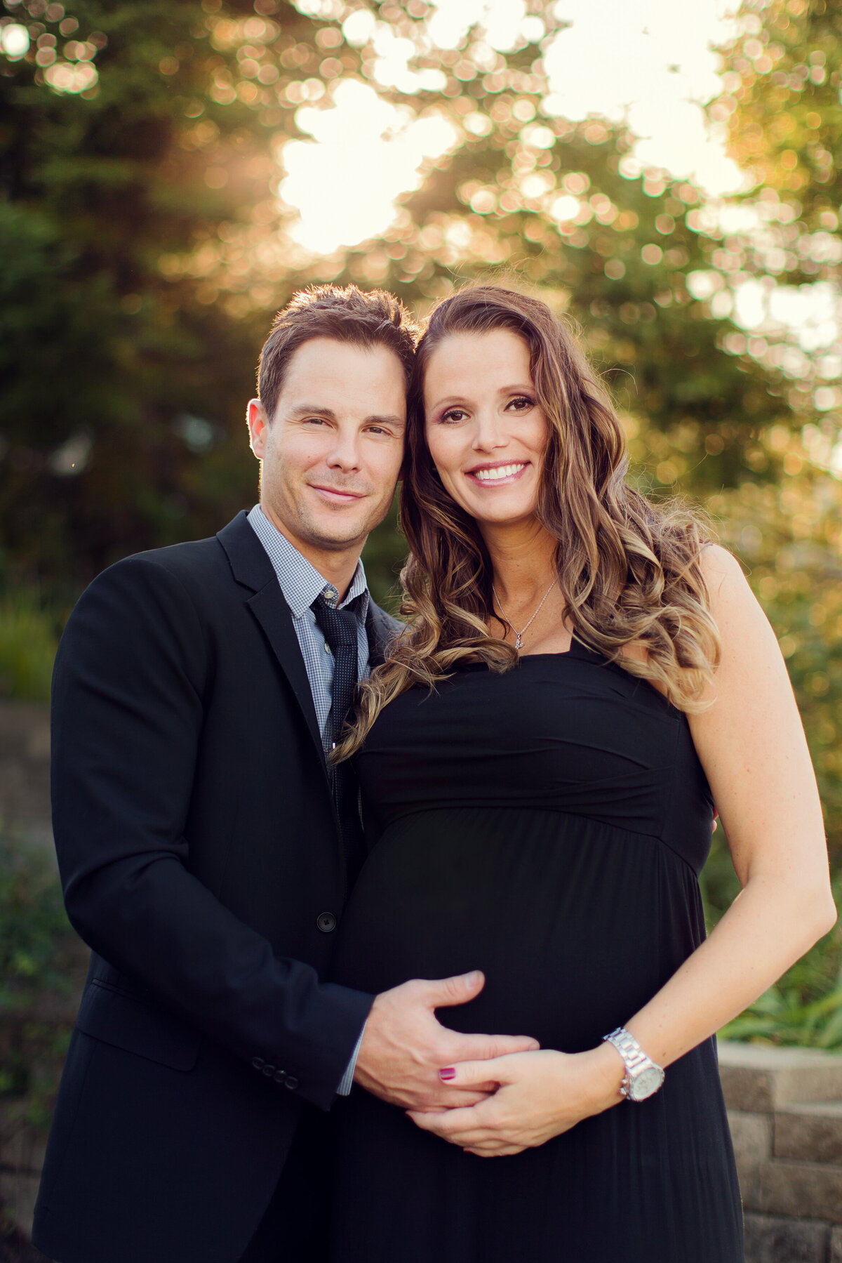 This husband in wife are dressed in a  black suit and black dress at sunset for a maternity session, their hands on her  belly.