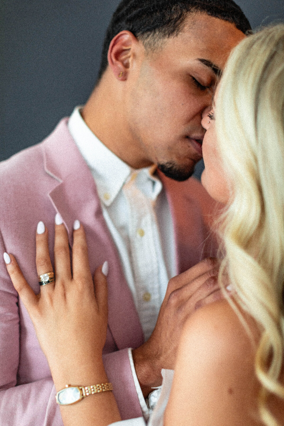 Close-up of a couple sharing an intimate moment, with the groom in a light pink suit and the bride's blonde hair cascading down. The soft lighting highlights their affection and the unique choice of a pastel-colored suit.