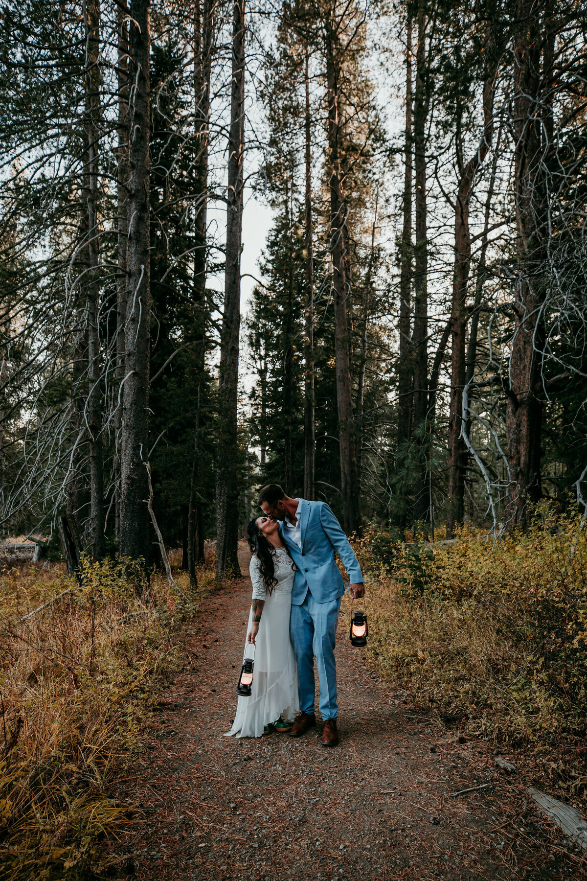 Bride and groom walking through a forested path at dusk, holding lanterns and sharing a quiet, intimate moment among the tall trees.