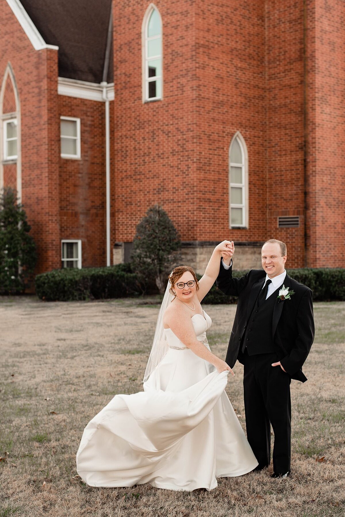 The bride and groom stand in front of a red brick church. The groom is wearing a black suit with a white shirt and black tie. He has one hand in his pocket and the other raised up to twirl the bride. The bride, holding the long train of her white satin dress, twirls back and forth. The bride is wearing a dress with a rhinestone belt and a strapless sweetheart neckline.
