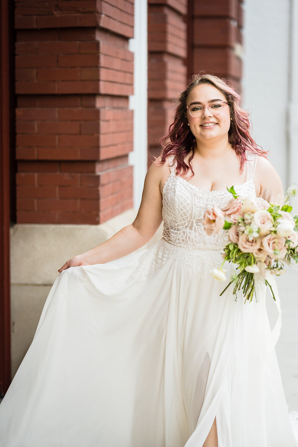 A marrier holds her bouquet in one hand and her dress in another and walks alongside the front of Fire Station One Boutique Hotel in Downtown Roanoke and smiles for a photo during their elopement in Virginia.