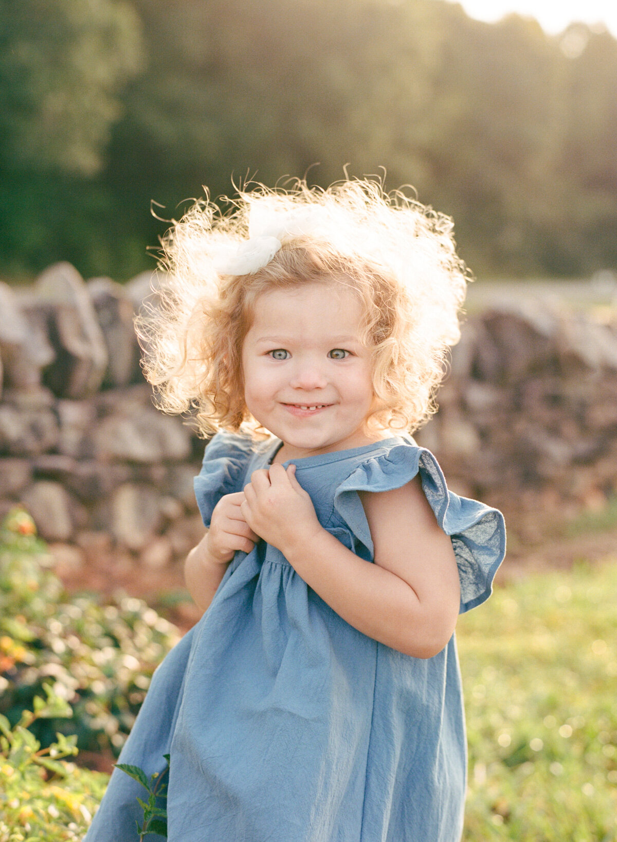 Toddler with bright blonde curls and blue eyes smiles directly at the camera while backlit by sun. Image by Raleigh maternity photographer A.J. Dunlap Photography.