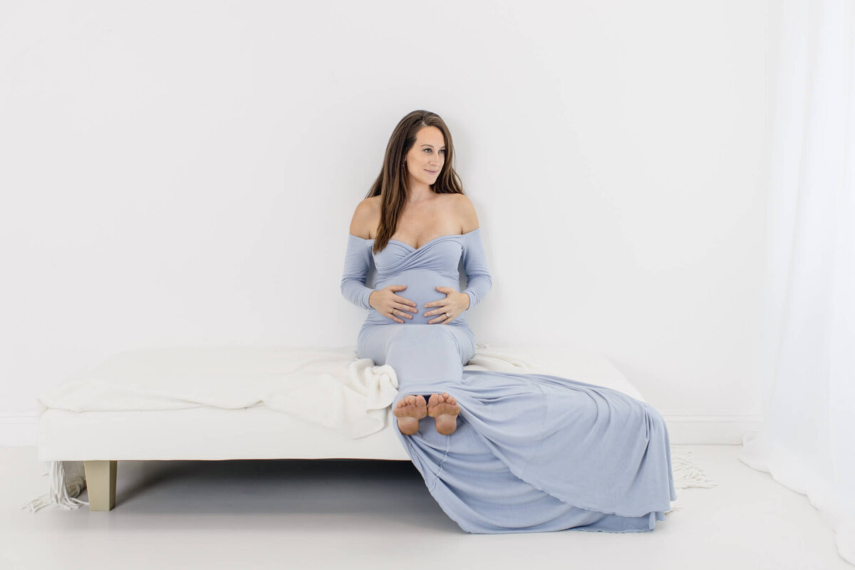 A pregnant woman in a flowing blue dress sits on a white bed against a minimalist white background. Her hands rest on her belly, and she gazes thoughtfully into the distance. The atmosphere is serene and peaceful.