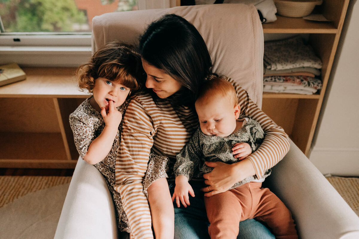 a young mother snuggles her young daughters in a light cream rocking chair
