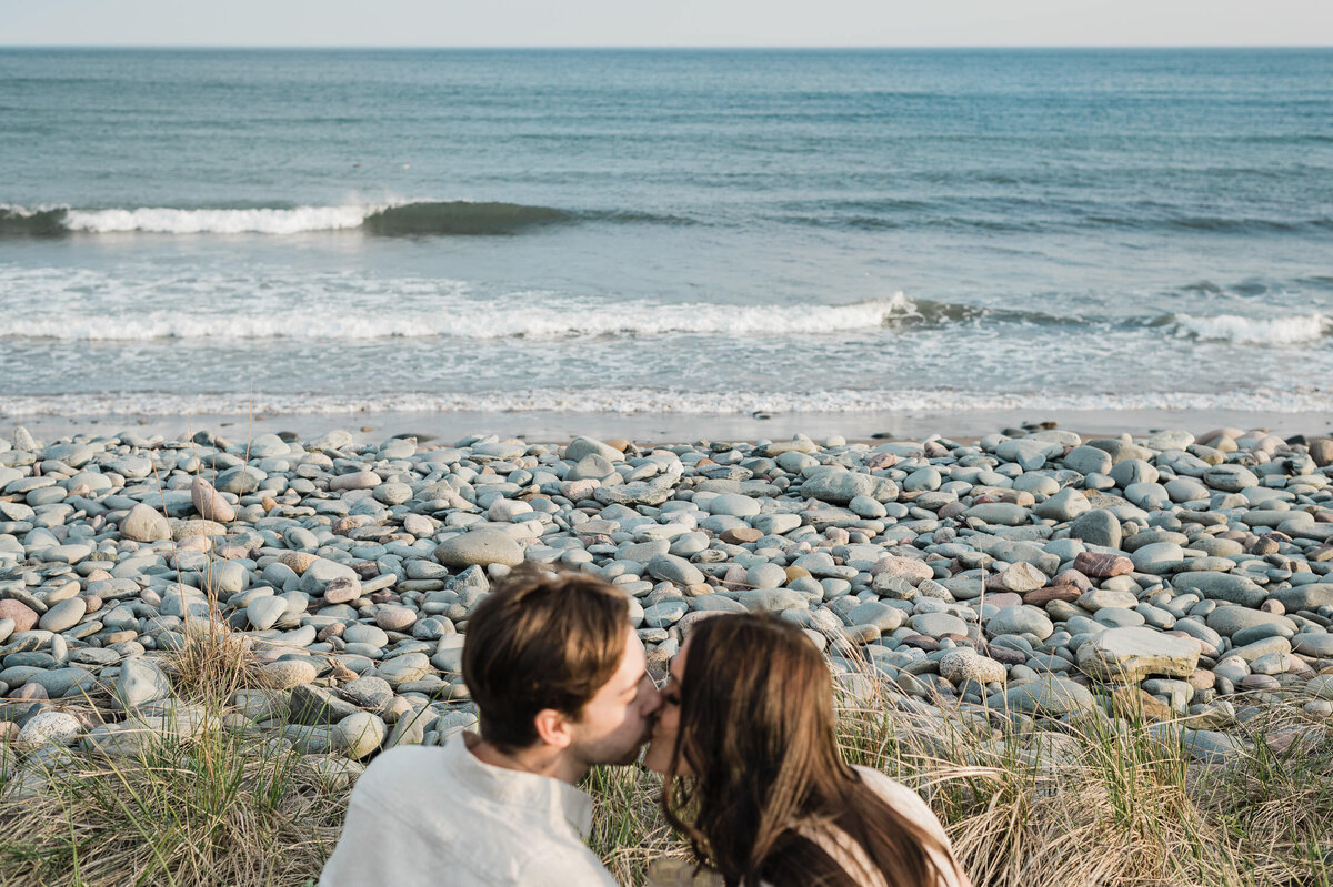 Couple kissing in front of rocky beach at sunset.