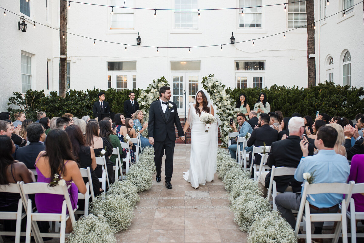 A bride and groom walk hand-in-hand down the aisle moments after saying "I do" during a beautiful outdoor ceremony at Casa Marina Hotel in Jacksonville Beach, Florida. Surrounded by lush white florals and rows of seated guests, the couple beams with joy under string lights and Mediterranean architecture. Captured by Heartful Films, a Jacksonville wedding photographer and videographer known for documenting timeless, emotion-filled celebrations with elegance and heart.