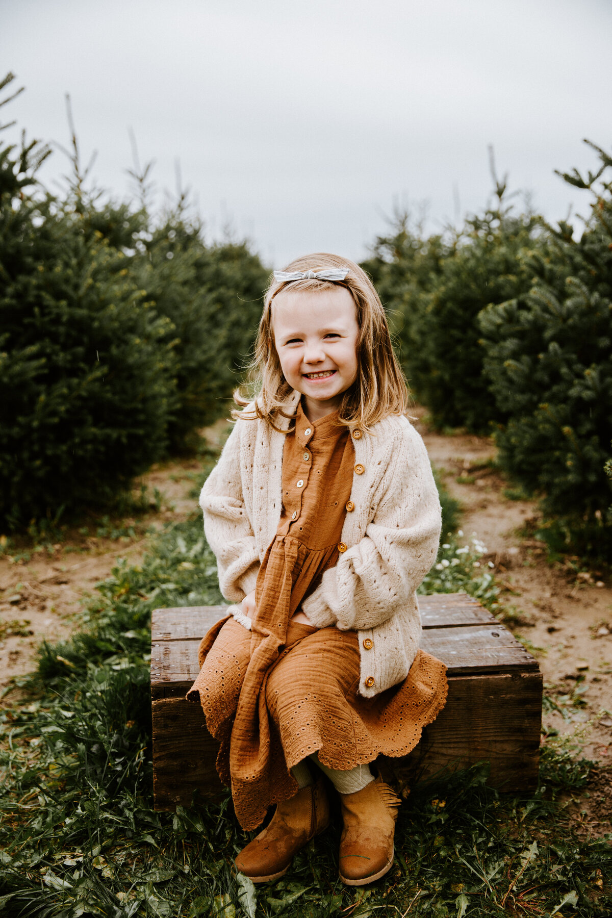 Tree Farm Christmas photos at Tree Lane Farms in London, Ontario. A young girl in a deep mustard dress and cream sweater is sitting on a crate amongst the Christmas evergreen trees with her hands in her pockets and smiling at the camera.