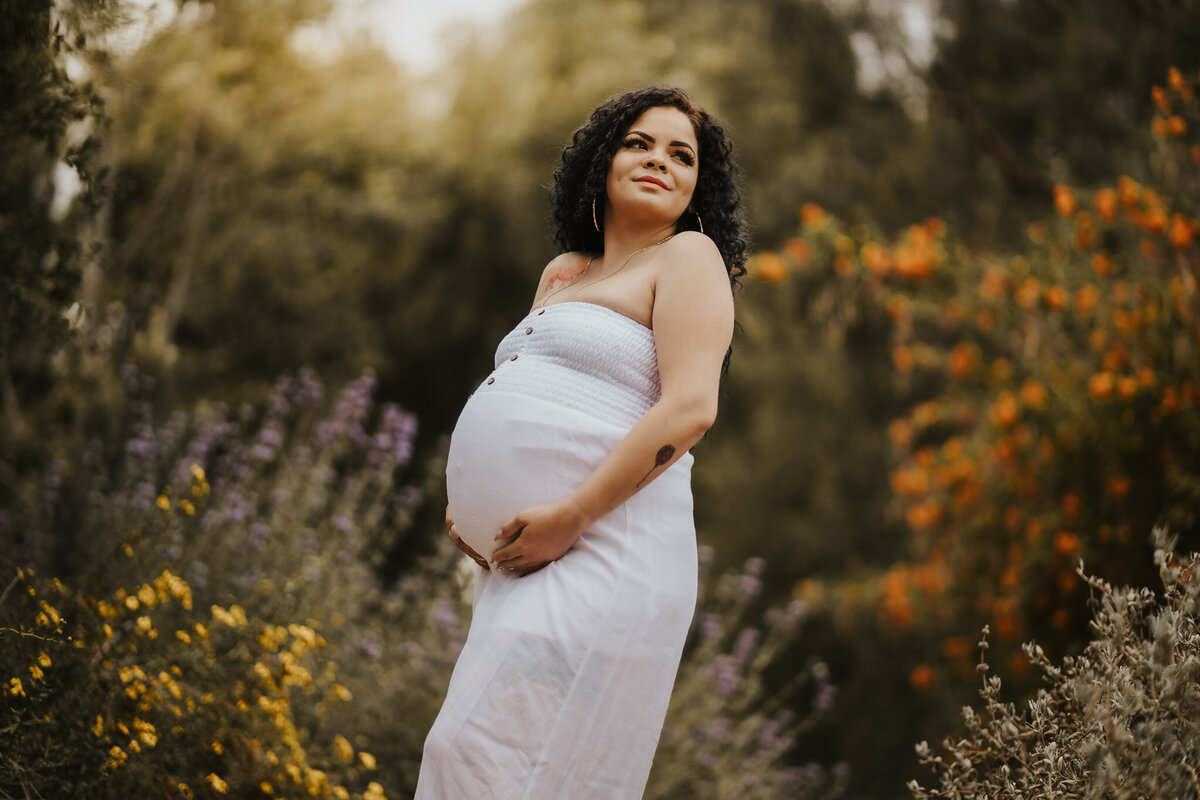 Expecting mom in a white dress holding her belly with green bushes and flowers in the background