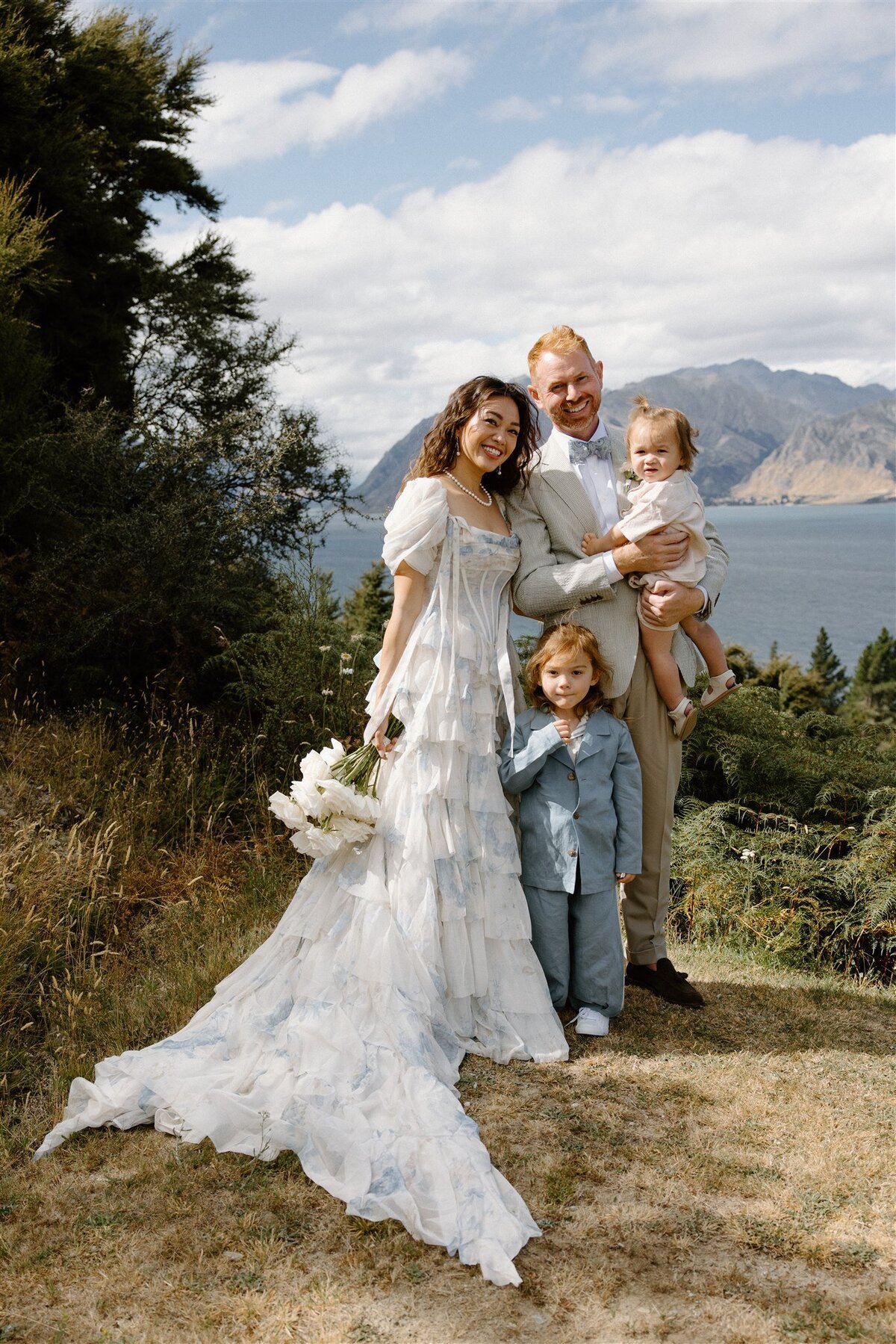 Wedding couple with their two children, including a toddler in the groom’s arms, sharing a happy moment with Lake Hāwea in the background.