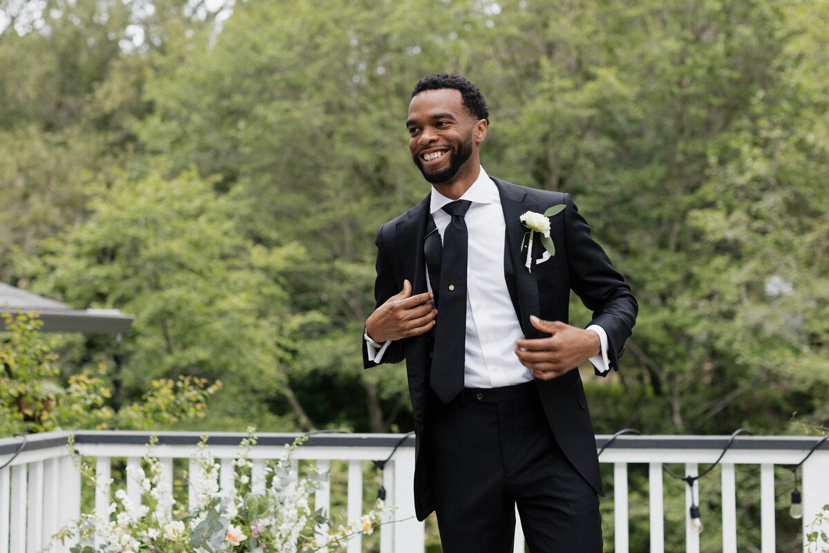 Candid wedding portrait of a groom adjusting his suit jacket and smiling before the ceremony. Captured outdoors with lush greenery in the background, this joyful moment showcases a modern groom's style with a classic black tuxedo and white boutonniere. Photographed by a luxury wedding photographer specializing in timeless and elegant wedding photography.