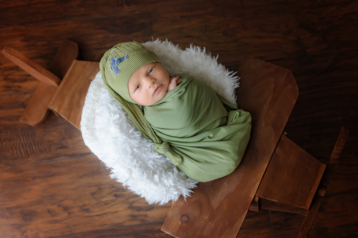 Newborn Photographer, a baby is swaddled in green sheets and wears a knit cap, sleeping on a fluffy blanket